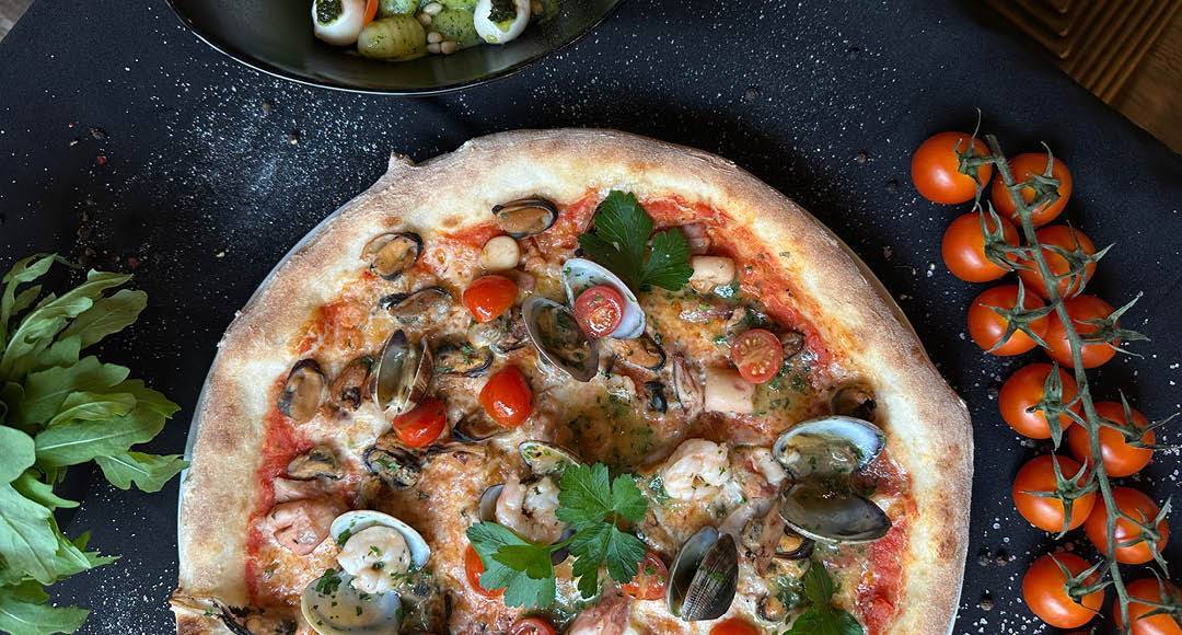La Osteria Italiener in Dresden Altstadt Pizza Meeresfrüchte Pizza mit Meeresfrüchten und danebenliegenden Rispen Minitomaten auf schwarzem Untergrund im La Osteria Italiener im Dresdner Zentrum